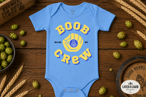 Blue baby onesie with 'Boob Crew' print on a wooden surface with hops and a barrel.