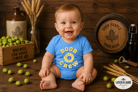 Baby wearing a blue onesie with 'Boob Crew' print, surrounded by beer-themed props on a wooden background.