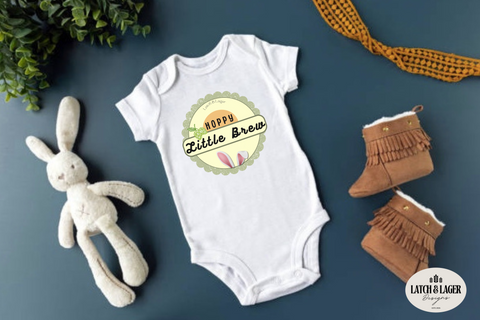 White baby onesie with 'Happy Little Brew' text, plush rabbit toy, and brown boots on a blue background.