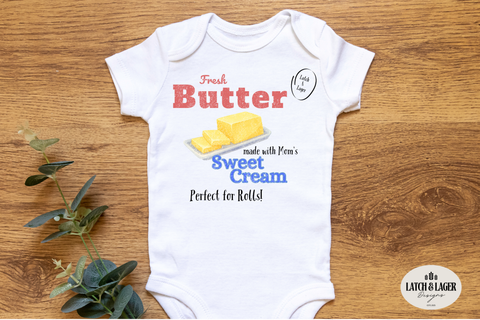 White baby onesie with 'Fresh Butter' text and graphics on a wooden surface