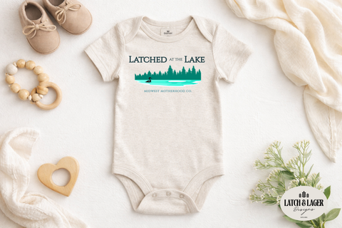 Baby onesie with 'Latched at the Lake' print on a white surface with toys and a blanket.