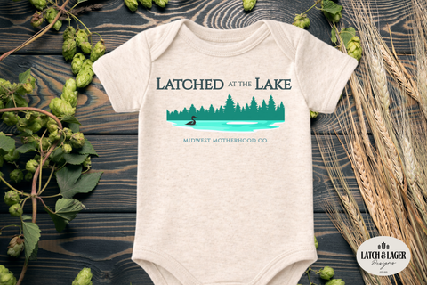 Beige baby onesie with 'Latched at the Lake' design on a wooden surface with greenery.