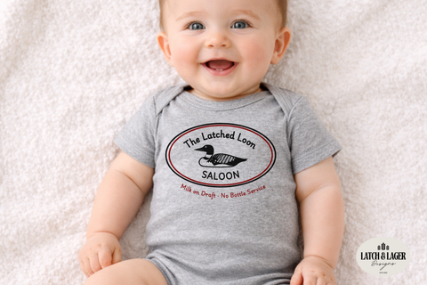 Baby wearing a gray onesie with a logo and text, sitting on a soft surface.