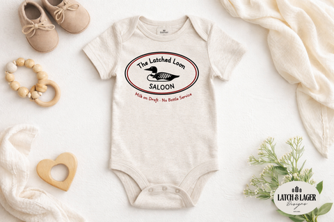 Baby onesie with 'The Latched Loom Saloon' design on a white surface with baby items.