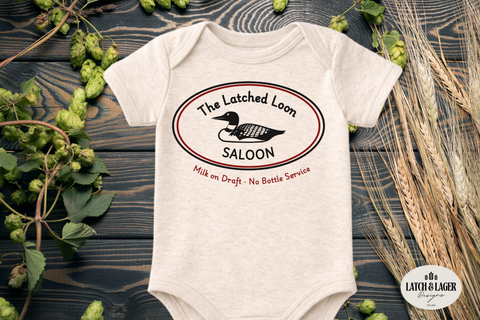 Baby onesie with 'The Latched Loon Saloon' design on a wooden surface with greenery.