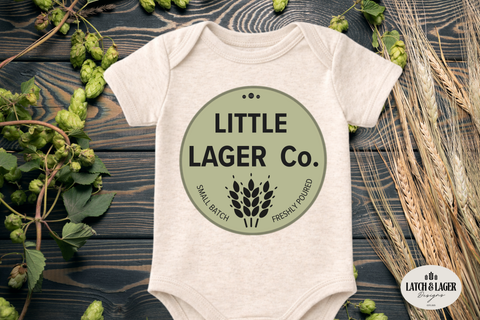 Baby onesie with 'Little Lager Co.' print on a wooden surface with hops and wheat.