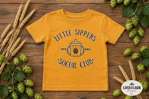 Yellow t-shirt with 'Little Sippers Social Club' design on a wooden background with hops and wheat.