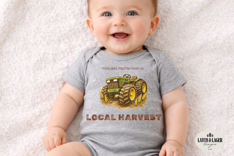 Baby wearing a gray onesie with a tractor design and text, sitting on a soft surface.