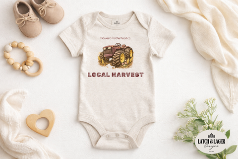 Baby onesie with 'Local Harvest' design on a white surface with toys and a blanket.