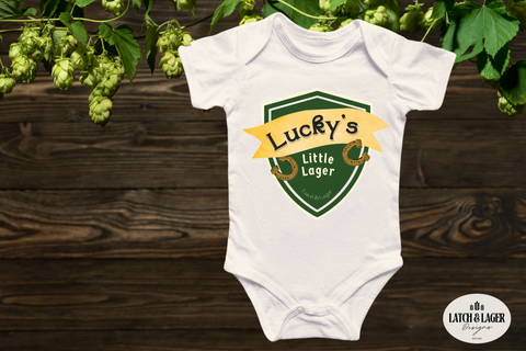 White baby onesie with 'Lucky's Little Lager' design on a wooden surface with hops in the background.