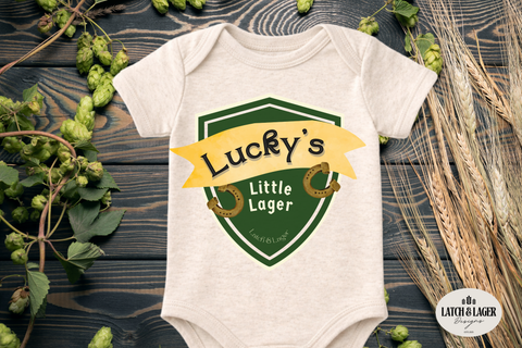 Baby onesie with 'Lucky's Little Lager' design on a wooden surface with greenery