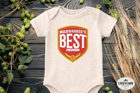 Baby onesie with 'Milkwaukee's Best' design on a wooden surface with hops and wheat.
