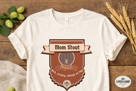 White t-shirt with 'Mom Stout' graphic design on a wooden surface with hops and wheat.