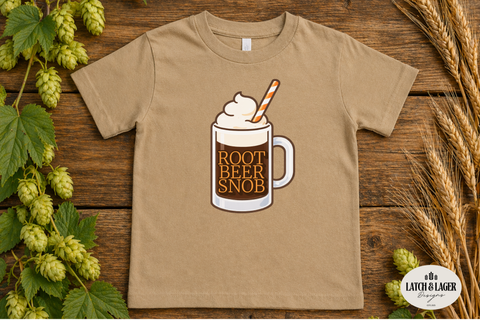 Beige t-shirt with root beer mug graphic on a wooden surface with hops and wheat.
