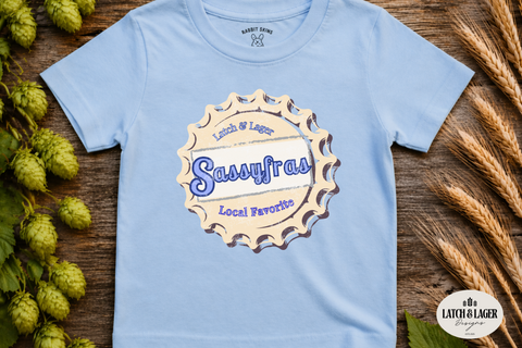 Light blue t-shirt with 'Sassyfras' logo on a wooden surface with hops and wheat.