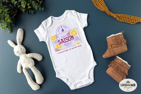 Baby onesie with 'Saison' design, plush rabbit toy, and brown boots on a blue surface.