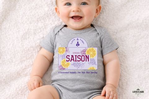 Baby wearing a gray onesie with a 'Latch & Lager' brand logo on a light background