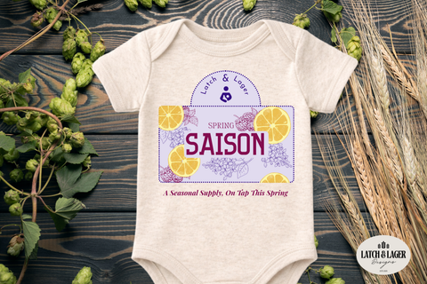Baby onesie with 'Spring Saison' design on a wooden surface with hops and barley.