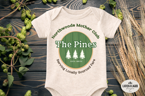Baby onesie with 'The Pines' design on a wooden surface with greenery