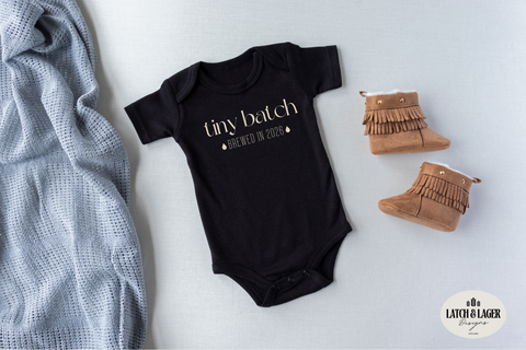 Black baby onesie with 'tiny batch' text, next to brown booties and a gray blanket on a light background.