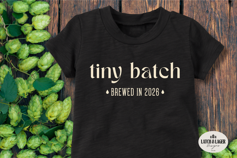 Black t-shirt with 'tiny batch brewed in 2028' text on a wooden background with hops.