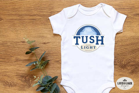 White baby onesie with 'TUSH Light' logo on a wooden surface with greenery.