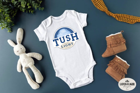 White baby onesie with 'TUSH LIGHT' logo, plush rabbit toy, and brown boots on a dark surface.